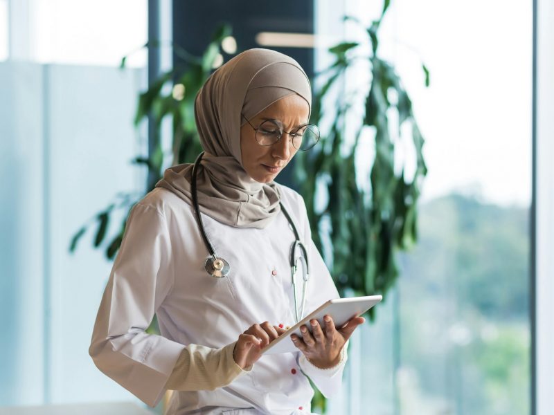 A worried and serious female doctor, an Arab, a Muslim is standing in the hospital, holding a